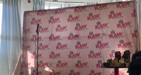 A pink backdrop with the repeated "RAW" logo, a microphone, and a small table with items in front, featuring a window and natural light.